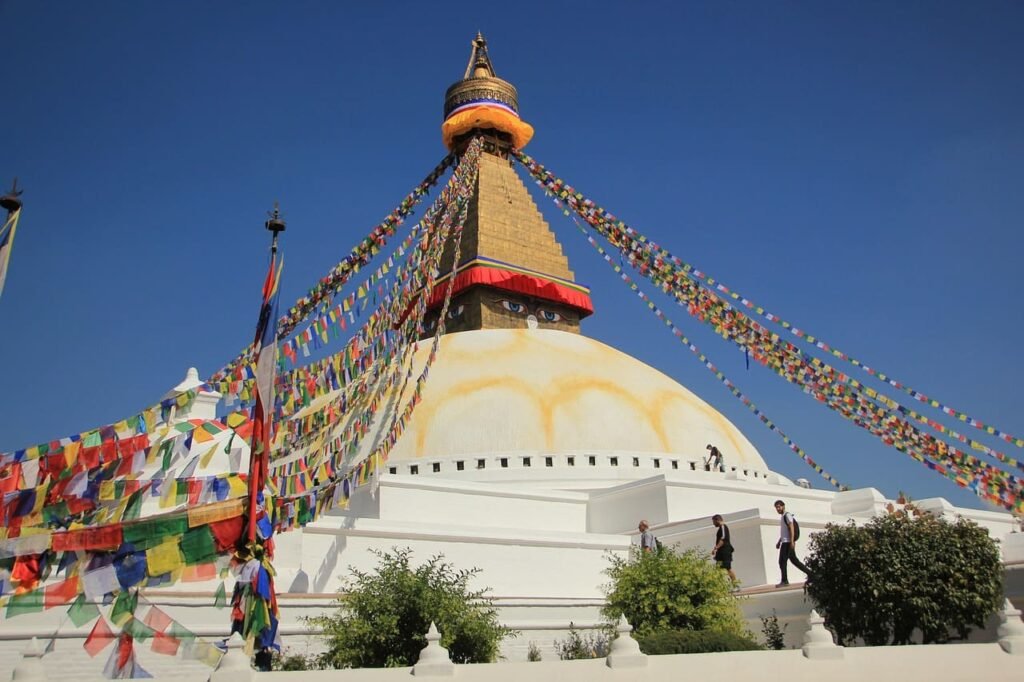 Boudhanath Stupa in Kathmandu, Nepal, with its iconic white dome and colorful prayer flags fluttering against a bright blue sky.